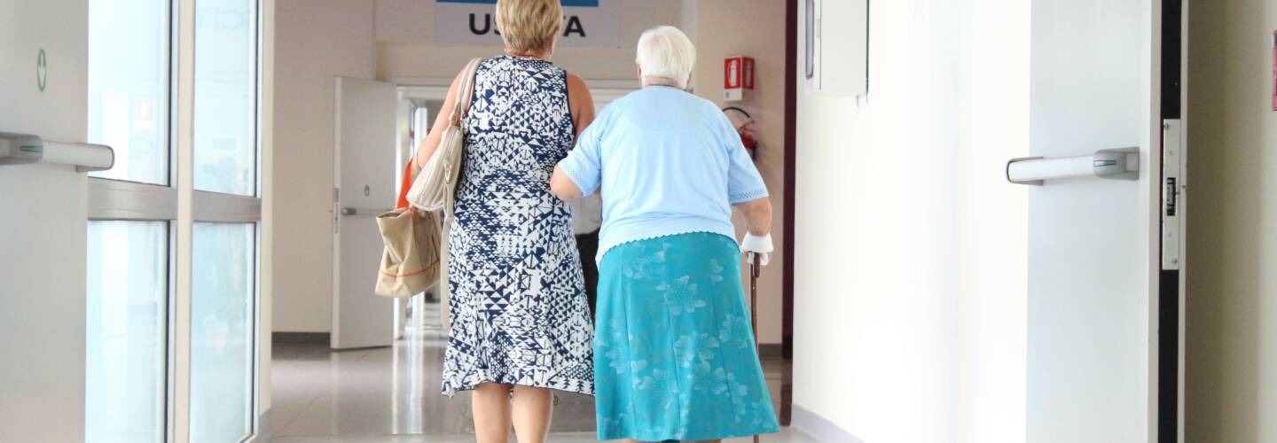 kaksi naista sairaalan käytävällä kävelee käsikkäin, toisella on kävelykeppi.