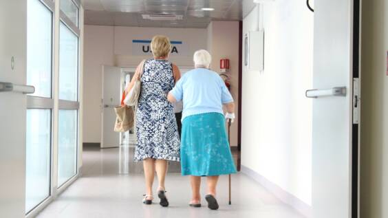 kaksi naista sairaalan käytävällä kävelee käsikkäin, toisella on kävelykeppi.