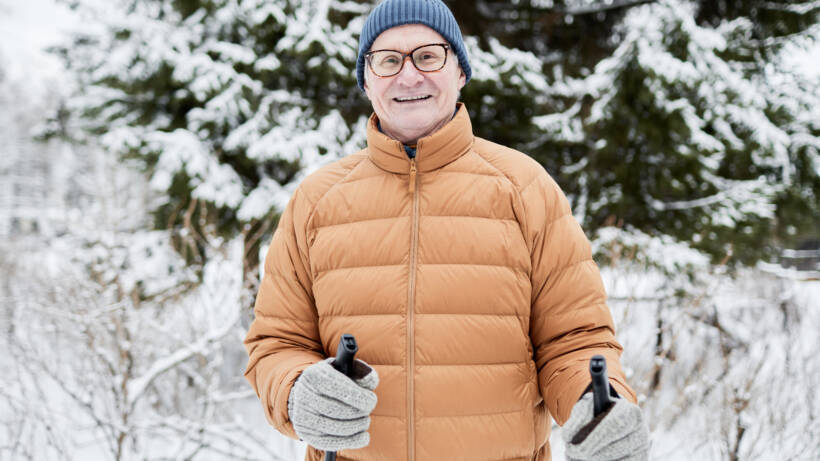 Seniorimies katsoo hymyillen kameraan sauvat kädessään. Taustalla näkyy talvista maisemaa.